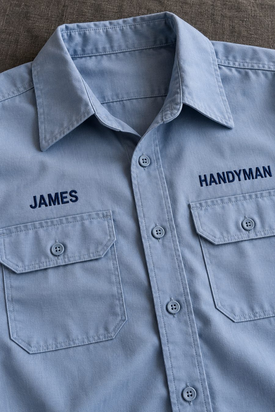 A light blue work shirt with JAMES embroidered above one pocket and HANDYMAN above the other in navy thread.