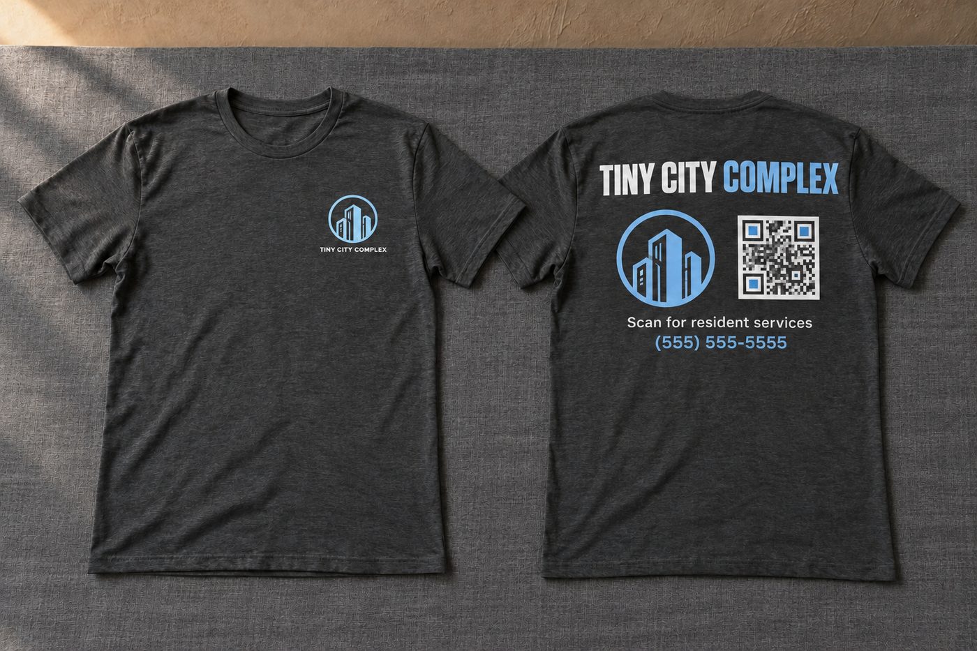 Two charcoal t-shirts side by side. The front shows a round building logo and TINY CITY COMPLEX on the chest. The back shows TINY CITY COMPLEX in large letters above the logo, a QR code and a phone number.